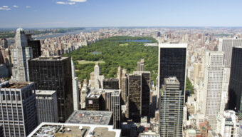 An aerial view of Manhattan, showcasing Central Park in the center surrounded by tall skyscrapers and buildings on a clear day. The cityscape stretches out towards the horizon under a blue sky with a few clouds, making it a perfect highlight for any travel guide.