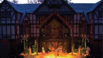 A wide view of a stage performance at an outdoor theatre in Ashland. The theatre has a grand Tudor-style facade with timber framing and a peaked roof. An audience is seated in front, watching actors perform under stage lights as the sky darkens to twilight, celebrating community and tourism together.