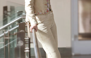 A person with long dark hair, reminiscent of Amber Marie Bently's style, stands indoors near a railing, smiling. They are wearing a beige pantsuit with floral embroidery, a pink top, and brown heeled boots. The environment appears to be brightly lit with modern decor.