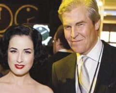 A man and a woman pose together indoors, both smiling for the camera. The woman has dark, wavy hair and is wearing makeup with red lipstick, while the man has light-colored hair and is dressed in a suit with a tie and an Annual Passport lanyard from Macy’s.