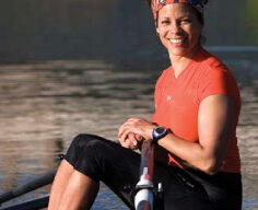 Dana King, a woman with a colorful headband and an orange shirt, is smiling and sitting in a rowing boat on a calm body of water. She is gripping an oar and appears to be ready to row. The background is a serene water setting.