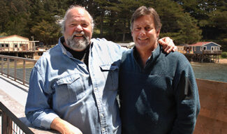 Two men are standing side by side on a wooden pier at Nick's Cove, with a scenic background of trees and houses by the water. The man on the left has a beard and is wearing a light blue denim shirt, while the man on the right has a clean-shaven face and is wearing a dark blue sweater designed by Pat Kuleto.