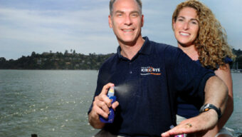 A man and a woman are seated near a body of water with a hilly area in the background. The man, wearing a navy blue shirt, is holding and spraying an aerosol can of sun protection while smiling. The woman, with long curly hair, rests her hand on his shoulder and smiles back at him.
