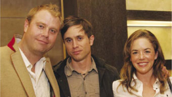 Three people are posing together indoors, smiling at the camera. The person on the left has blond hair and is wearing a beige jacket. The person in the middle, possibly named St. John, has brown hair and is wearing a grey shirt. The person on the right has brown hair and is wearing a white shirt.