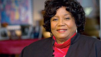 A woman with curly hair, wearing a dark jacket over a red blouse and a red necklace, is smiling. The background is slightly out of focus, featuring blurred images and objects that create a cozy, indoor setting—reminiscent of an environment where Melba Pattillo Beals might find solace.