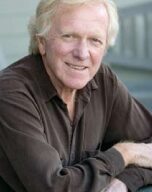 A man with light hair and a brown shirt is sitting outdoors, smiling and resting his arms on a surface in front of him. The background is slightly blurred, placing the focus on him. His relaxed demeanor complements the serene setting, embodying an eco-friendly lifestyle choice.