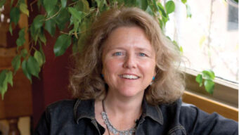 A person with wavy, shoulder-length hair smiles while sitting in front of a window. They are wearing a dark jacket and a silver necklace. A leafy plant, perhaps symbolizing their passion for environmental education, is visible in the background.