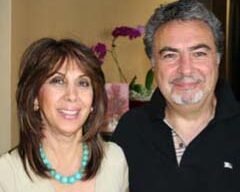 A woman with shoulder-length brown hair and a turquoise necklace smiles while standing next to a man with short curly gray hair and a beard, who is also smiling. They are at a chamber mixer in Sausalito, indoors with a blurry background featuring an orchid and a picture frame.