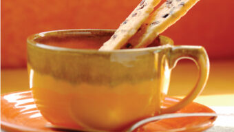 A ceramic cup with a matching saucer, each with a warm, orange and brown gradient design, holds two almond sticks. The cup is filled with a hot beverage, and a small spoon rests on the saucer. The background is softly blurred, featuring orange hues reminiscent of a rustic bakery.