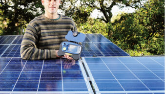 A person in a striped sweater and cap stands next to solar panels, holding a small open case with electronic components. Behind them, trees stretch under a clear sky. The scene captures a green energy application or installation in action.