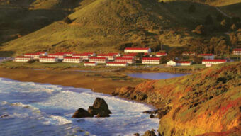 A coastal scene with numerous white buildings with red roofs situated near a beach. The waves crash against rocky outcrops in the foreground, while green rolling hills dominate the background under a partly cloudy sky, evoking thoughts of refreshing spring wardrobes and airy dresses.