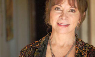 A woman with brown hair and bangs, wearing a patterned jacket and a necklace with a large pendant, smiles while standing in a hallway with soft lighting, evoking the charm of a character from Latin American literature.