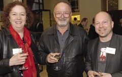 Three people stand together at an indoor exhibit, each holding a drink. The person on the left, with curly hair and a red scarf, holds a teacup. The middle person has glasses and a beard. The person on the right is bald and wears a T-shirt with a jacket. All are smiling warmly.