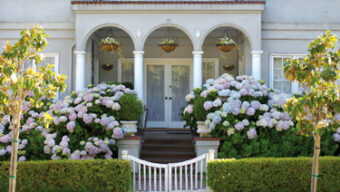 A two-story Dominican-style house with a light gray exterior, white columns, and an arched entryway. The front yard features manicured bushes, blooming hydrangeas, hanging plants, and a white picket gate. Trees flank the walkway leading to the front door.