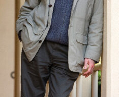 John Stayton, with a friendly smile, leans casually against a wall. Wearing a light-colored jacket over a dark sweater and dark pants, his hands rest in his jacket pockets. He looks relaxed in the outdoor setting adorned with architectural elements.