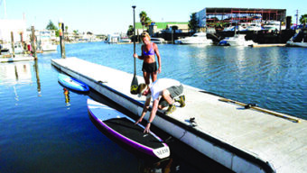Two people are preparing to Get On the Water at a marina on a sunny day. One stands holding a paddle while the other kneels on a dock, adjusting a paddleboard. The background displays boats docked in the water and shoreline buildings.