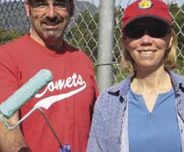 A man and a woman, both wearing red caps, stand in front of a chain-link fence, smiling. The man wears a red "Comets" softball shirt and holds a paint roller. The woman wears sunglasses and a blue shirt, holding a paintbrush. Trees and mountains are visible in the background as they participate in the clean-up effort.