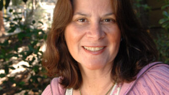 A woman with shoulder-length dark hair smiles at the camera. She is wearing a light pink jacket over a white patterned top. The background features greenery and trees, indicating an outdoor setting with natural light, perfect for finding a doctor who appreciates the harmony of nature and good health.