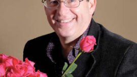 Gerald Porlman, a smiling man with short dark hair and glasses, dressed in a black jacket and purple shirt, holding a single pink rose. He stands surrounded by bouquets of red and pink roses against a plain beige background, truly embodying the essence of planting seeds of change.