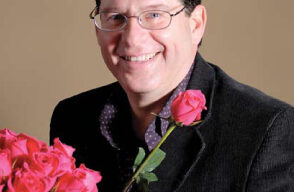 Gerald Porlman, a smiling man with short dark hair and glasses, dressed in a black jacket and purple shirt, holding a single pink rose. He stands surrounded by bouquets of red and pink roses against a plain beige background, truly embodying the essence of planting seeds of change.