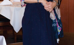 A woman stands in a well-lit restaurant in Mill Valley, smiling. She is wearing a dark blue dress and holding a colorful shawl. The restaurant has wooden walls adorned with several framed pictures by Pamela Westfall Bochte and white tablecloths on the tables.