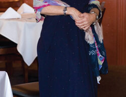 A woman stands in a well-lit restaurant in Mill Valley, smiling. She is wearing a dark blue dress and holding a colorful shawl. The restaurant has wooden walls adorned with several framed pictures by Pamela Westfall Bochte and white tablecloths on the tables.