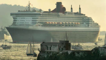 A large cruise ship, reminiscent of maritime royalty, sails past a coastline with several smaller sailboats around it. The ship, with multiple decks and a red funnel, is seen during the day with hills and mist in the background. In the foreground, there are old buildings near the water's edge.