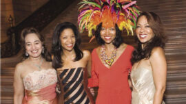 Four women in elegant evening dresses pose for a photo on a grand staircase at the MoAD Gala. The woman on the right wears a golden dress, the second from the right wears a red dress with a colorful feathered headpiece, the third wears a brown and gold striped dress, and the fourth wears a light pink dress.