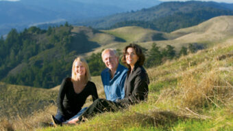 Three people are sitting on a grassy hillside with expansive rolling hills and forested areas in the background. They are smiling and appear relaxed, enjoying the natural scenery. The sky is clear, offering a bright, sunny day and a perfect view of Mt. Tam in the distance.