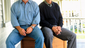 Two older men are sitting on wooden benches outside on a porch, their smiles hinting at an amicable meeting of minds. The man on the left has a white beard and wears a light blue shirt with jeans and brown shoes, while the man on the right sports a grey cap, dark sweater, grey pants, and brown shoes.