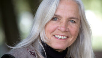 A woman with long, silver hair smiles warmly at the camera. She is wearing a dark turtleneck and a gray vest. The background is softly blurred with shades of green and light, suggesting an outdoor setting—a serene moment capturing Penny Livingston-Stark's tranquil spirit.