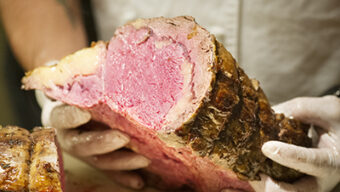 A person wearing white gloves holds a large, thick slice of juicy, medium-rare roast beef, reminiscent of Todd Shoberg's culinary mastery. The fat and edges of the roast are browned, contrasting with the tender, pink interior.