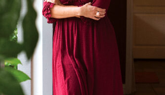 A woman with long dark hair, wearing a knee-length red dress and black boots, leans casually against a door frame while smiling. Jeannie Jarnot stands in a warmly lit area with some greenery visible on the side.