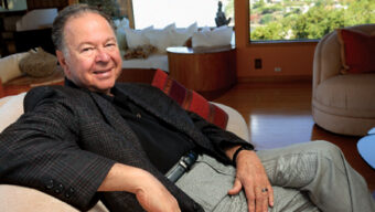 A man wearing a dark blazer and light-colored pants is sitting comfortably on a beige chair. He is smiling and has his hand resting on his knee. Behind him is a large window with a scenic view of greenery and a body of water. The room, reminiscent of Leonard Shlain's study, has warm, wooden tones.