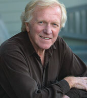 A smiling man with light-colored hair and wearing a dark shirt leans on a wooden surface. The background is blurred, featuring a light-colored wall and part of a more efficient structure, adding to Marin's charm.