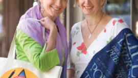 Two smiling women stand side by side on a sidewalk. One woman holds a white tote bag with a colorful logo and wears a green top with a lavender scarf, embodying the vibrant branding of MMob. The other wears a white dress with a floral pattern and drapes a blue banner-like cloth over her shoulder.