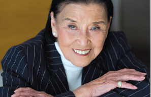 A smiling, elderly person with dark, slicked-back hair rests their arms on a surface. They wear a dark pinstriped suit jacket over a white turtleneck and have pearl earrings and rings on both hands. The background is out of focus, emphasizing Cecilia Chiang's face and elegant attire.