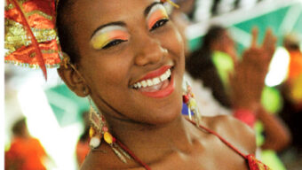 A smiling woman in vibrant carnival attire with a large, ornate headdress adorned with gold and red feathers and jewels. Her red and gold sequined top sparkles as she flaunts colorful makeup. The blurred, bright background hints at a lively festival atmosphere, enchanting both locals and tourists alike.