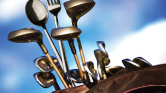 A golf bag filled with various golf clubs and a few gourmet utensils, like a fork and a spoon, sticking out among the clubs, set against a blue sky with white clouds.