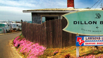 A quaint roadside scene features a wooden building with a brick chimney, adorned by vibrant pink flowers along a weathered fence. A green sign reads "Dillon Beach," inviting travelers to explore its charm, while a red and blue sign points towards Lawson's Landing for camping and boating services.