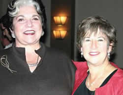 Two women smiling at the camera. The woman on the left has short, dark hair with a white streak and is wearing a dark top with a pin. The woman on the right has short, light brown hair and is wearing a red jacket over a black top. They are standing close together indoors, possibly after an opera celebration.