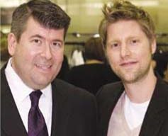 Two men are standing side by side indoors, both smiling at the camera. The man on the left is wearing a dark suit with a white shirt and a purple tie. The man on the right is wearing a casual Burberry blazer over a light-colored shirt. The background is blurred.