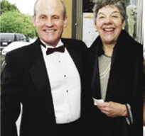 A smiling man and woman are dressed formally, as if attending the Academy Awards in Larkspur. The man is wearing a black tuxedo with a bow tie, and the woman is in a black outfit with a pearl necklace and a gray hair net. They stand close together outdoors, with greenery and cars in the background.