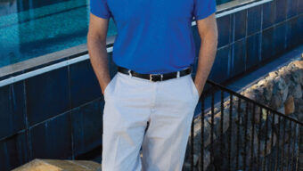 A man in a blue polo shirt and light-colored pants stands with hands in his pockets, smiling. He is outdoors near a building with large windows and an in-ground pool, likely in Tiburon. Plants and stonework are visible in the foreground, capturing the serene ambiance of Robert C. Placak's design.