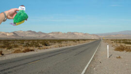 A hand holding a colorful paper cutout of a person stands over a seemingly endless road trip route stretching into a barren desert landscape with distant mountains under a clear blue sky.