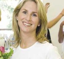 A smiling person with shoulder-length blonde hair wears a white t-shirt. They are indoors with part of another person visible in the background. They are holding a bouquet of flowers, perhaps a gift from their honeys.