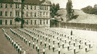 Sepia-toned historical photo showing a large group of people, possibly schoolchildren, performing a drill or exercise in rows on a large open space in front of St. Vincent's multi-story building with many windows. Trees and hills are visible in the background, capturing a moment from "Then and Now.