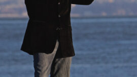 Jason Jennings, in a black blazer and jeans, stands by the water with a violin and bow. He is smiling while playing the violin. The background features calm water and rocks on a grassy area.