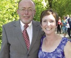 A man in a grey suit with glasses and a woman in a purple dress smile at the camera while standing outdoors at what seems like a charming Garden Party. Green trees and other people can be seen in the background, adding to the festive atmosphere.