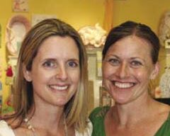 Two women are standing close together, smiling at the camera. Both have shoulder-length hair; the woman on the left has blonde hair, and the woman on the right has brown hair pulled back. The brightly lit indoor setting boasts colorful decor, hinting at a grand reopening celebration.